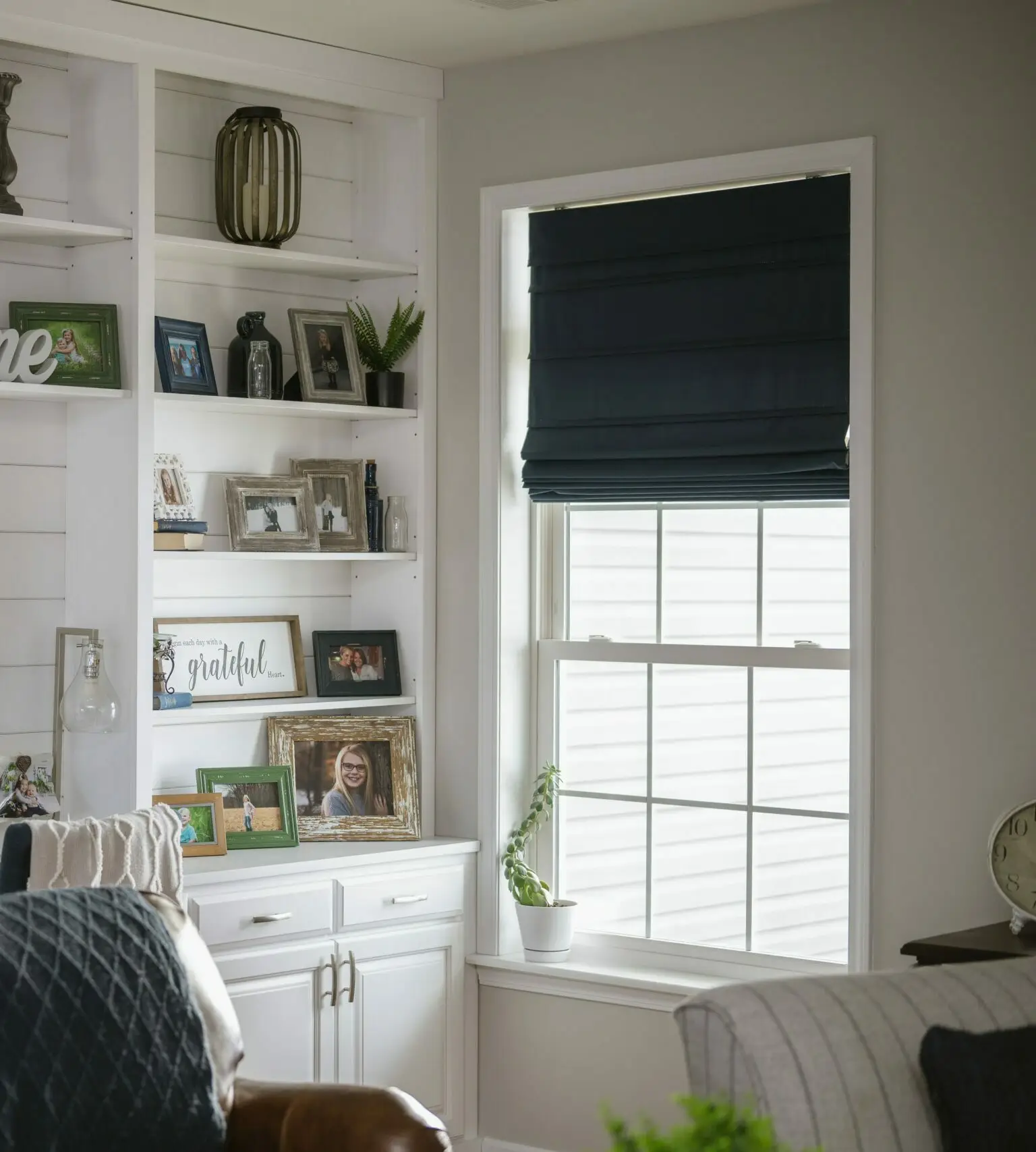 White built-in bookshelf with family photos, frames, a small plant, and decorative items beside a window with a dark roller blind